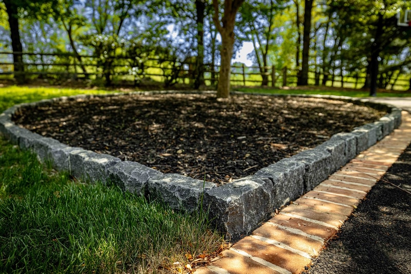 Low-angle view of the Hockessin entry courtyard: a small brass path lamp anchors the foreground, the brick paver courtyard sweeps to the arched front door beyond, with a mulched bed of small shrubs and a stone-edged border framing the right