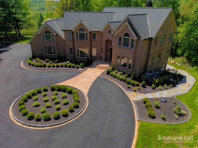 Three-quarter angled view of the Hockessin estate front: the multi-gable brick home with arched windows centered behind the curved brick paver driveway loop, the boxwood-planted oval centerpiece with rows of mounded boxwoods, and the brick paver entry walkway leading to the front doors, framed by mature trees and rolling lawn
