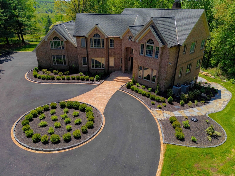Aerial three-quarter view of a Hockessin Delaware estate: a multi-gable brick home sits at center, fronted by a sweeping curved brick paver driveway that wraps around a circular boxwood-planted oval centerpiece, with mature trees framing the property and a paver entry courtyard at the front door