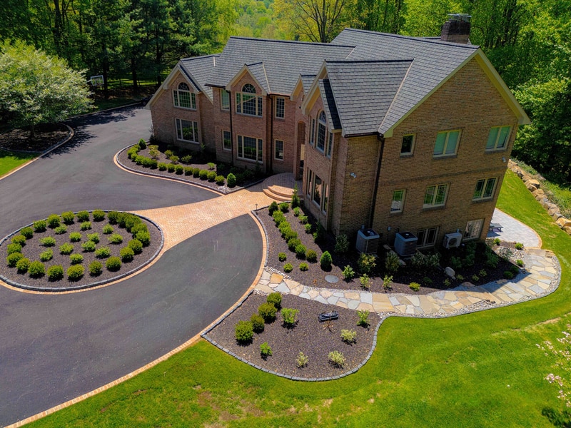 Aerial three-quarter view of a Hockessin Delaware estate from the side: the multi-gable brick home fills the right of the frame, with the curved paver-bordered driveway and circular boxwood-planted oval centerpiece in front, a side flagstone walkway threading past the equipment area, and mature trees framing the property
