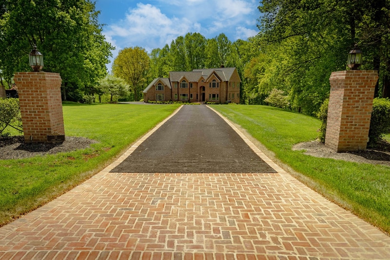 Long view up the Hockessin driveway from the road: a brick paver runner stretches between two brick gate columns with lantern caps, a smooth lawn extends on both sides, and the brick estate sits centered at the end of the approach
