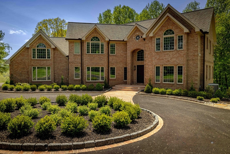 Low-angle ground-level view along the Hockessin driveway: the curved brick-edged asphalt drive sweeps across the foreground toward the brick estate, with dramatic blue-and-white cumulus clouds filling the sky overhead