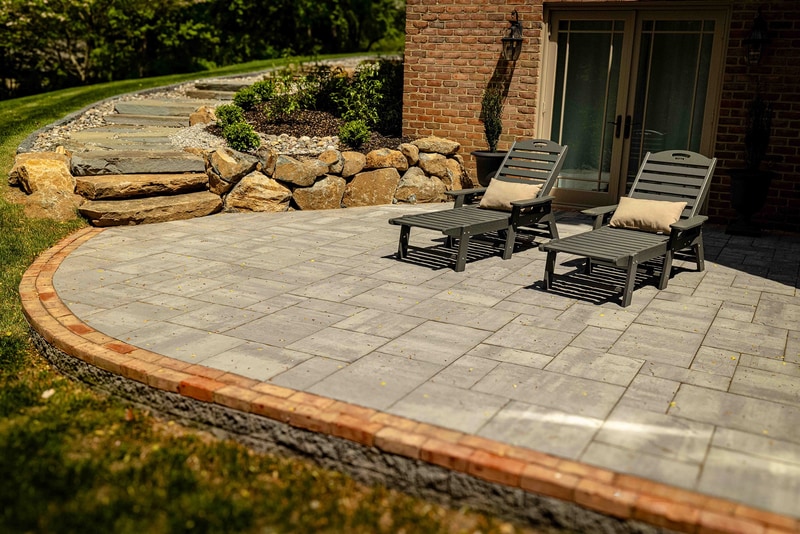 Detail view of a Hockessin planting bed edge: a low stacked-stone wall in dark gray fieldstone separates a mulched bed of mounded boxwoods from the asphalt drive, with a brick paver runner curving past at the right edge