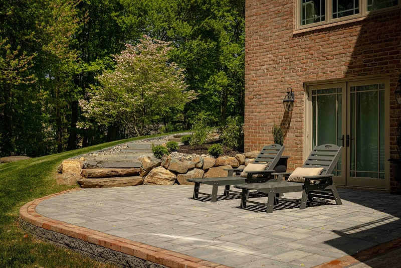 Eye-level view of the Hockessin back patio: a pair of dark wood chaise lounges with cream cushions sit on a curved gray paver patio with a brick band edge, with a dogwood in bloom and a tumbled stone retaining wall behind, and a smooth lawn beyond
