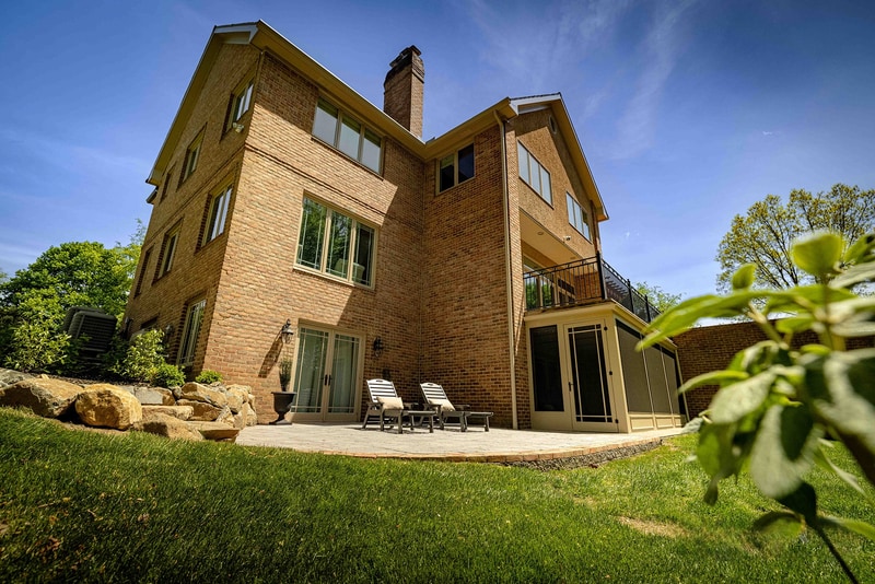 Wide ground-level view of the back of the Hockessin estate: the tall brick facade with chimney rises across the frame, a small back patio with a pair of chaise lounges sits below the walkout level, and a smooth green lawn extends to the woodland edge at the right