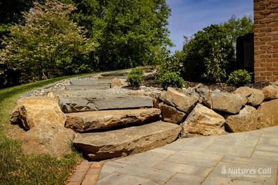 Wide view of the Hockessin round back patio: a circular gray paver pad with a brick band edge holds two dark wood chaise lounges, ringed by large rough boulders and a wire-cage gabion edge, with the home's tall brick wall on the right and a sloping lawn beyond