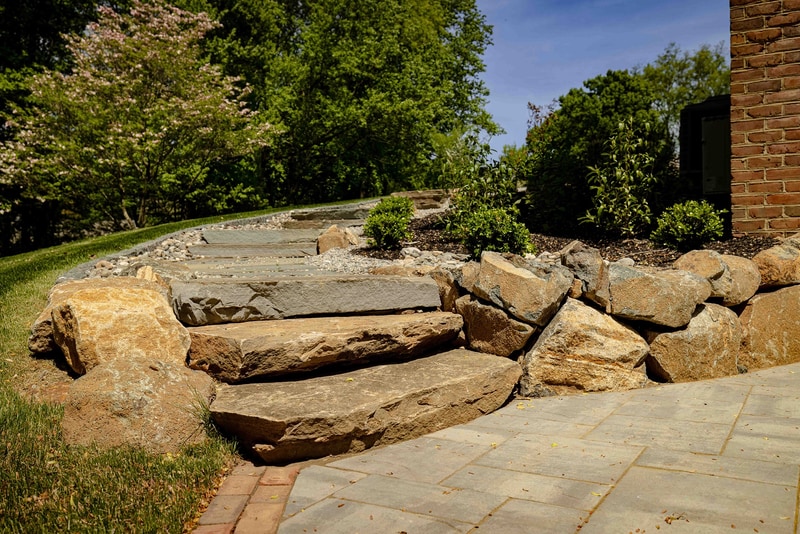 Wide view of the Hockessin round back patio: a circular gray paver pad with a brick band edge holds two dark wood chaise lounges, ringed by large rough boulders and a wire-cage gabion edge, with the home's tall brick wall on the right and a sloping lawn beyond