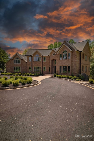 Cinematic portrait of the Hockessin home at sunset: the brick estate facade glows in late evening light beneath a dramatic orange-and-blue cloud-streaked sky, with the curved paver driveway sweeping in from the foreground