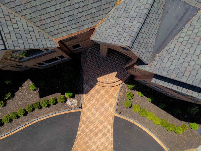 Close angled aerial view of the Hockessin home's roofline and front entry: the multi-gable slate-style roof frames a brick paver entry courtyard below, where the curved paver driveway meets the front doors in a graphic geometric composition