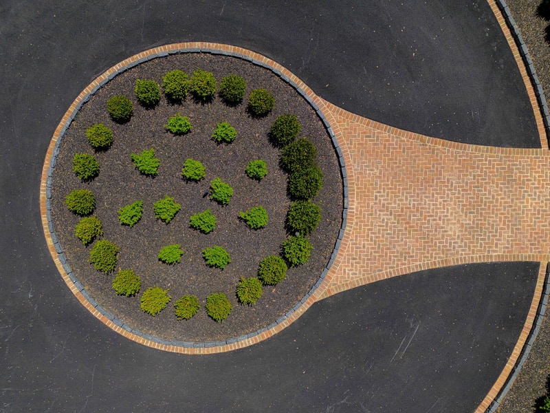 Top-down drone view of a Hockessin estate boxwood-planted oval centerpiece: a perfect oval bed of mulch ringed by a brick paver border holds rows of round mounded boxwoods at the heart of the curved paver driveway loop — Nature's Call landscape and hardscape project