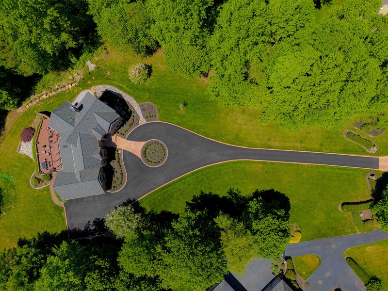 Drone view straight down on the Hockessin driveway: the long asphalt drive flares into a circular paver loop at the house, the boxwood-planted oval centered inside the loop, with mature trees flanking the approach