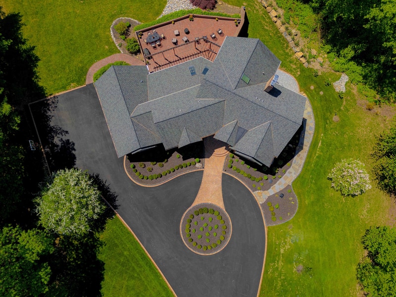 Drone view straight down on the back of the Hockessin estate: the multi-gable roof plan is visible at the bottom, a circular paver patio with chaise seating sits at the top of the frame, and the curved driveway sweeps in from the right