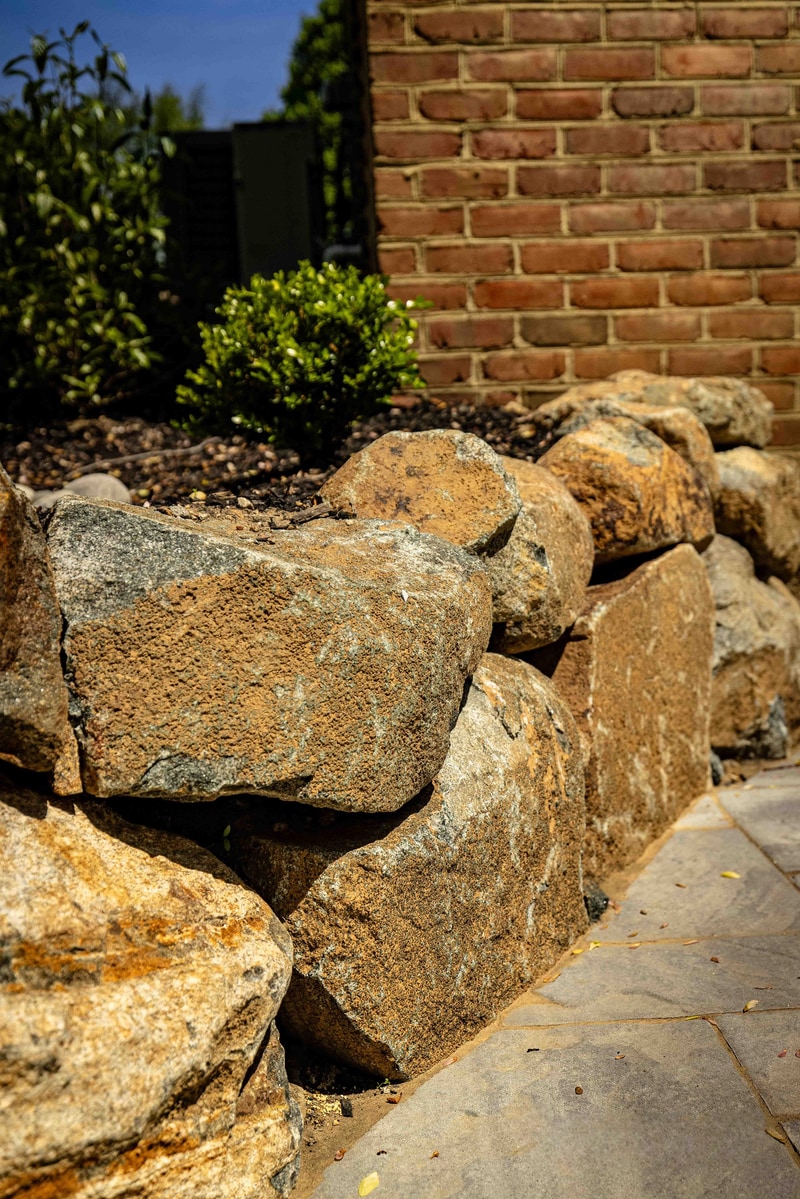 Portrait detail of a Hockessin boulder retaining wall: large rough fieldstone boulders are dry-stacked against a brick exterior wall, with a single mounded boxwood at the top of the wall and a small mulched bed running alongside the bluestone path at the base