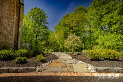 Stone stepping-stone pathway winds through mulch beds with ornamental shrubs framing a wooded backdrop in Hockessin, DE residential landscape.