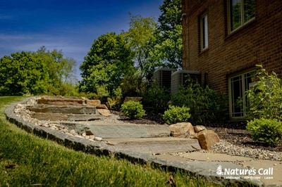 Curved stone retaining wall with flagstone patio adjoins brick home in Hockessin, DE, surrounded by shrub plantings and lawn backdrop.