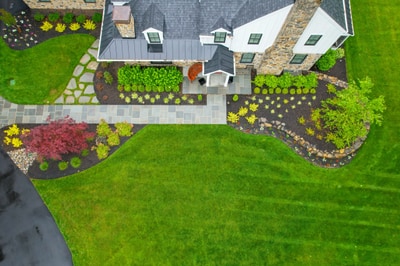 Aerial view of a manicured landscape with yellow flowering beds, burgundy foliage, paver walkways, and expansive green lawn surrounding a contemporary home.