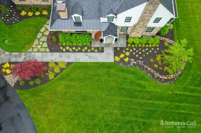 Aerial view of a manicured landscape with yellow flowering beds, burgundy foliage, paver walkways, and expansive green lawn surrounding a contemporary home.