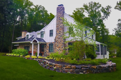 Contemporary farmhouse with stone chimney features foundation plantings of purple flowers, ornamental grasses, and a curved stone retaining wall set in green lawn.