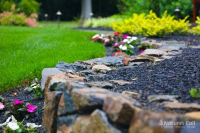 Stone edging defines flower beds with vibrant pink and white blooms, yellow shrubs, and mulch, bordered by a manicured lawn.