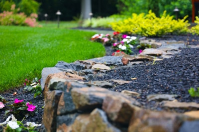 Stone edging defines flower beds with vibrant pink and white blooms, yellow shrubs, and mulch, bordered by a manicured lawn.