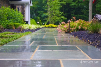 Slate paver walkway with brass inlays leads through perennial beds with pink and yellow flowers, ornamental grasses, and mature plantings flanking a home.