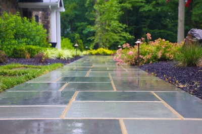 Slate paver walkway with brass inlays leads through perennial beds with pink and yellow flowers, ornamental grasses, and mature plantings flanking a home.