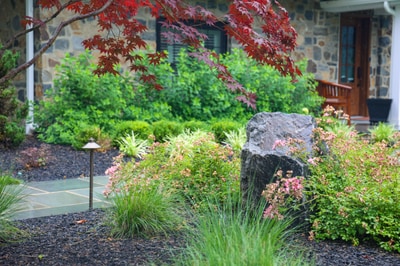 Modern landscape bed with black mulch, ornamental grasses, pink flowering shrubs, and weathered stone accents near a stone home.