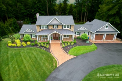 Aerial three-quarter view of the front of the North Wilmington home: the stone-and-cedar-shake facade sits centered, the curved tan paver driveway with charcoal Belgium-block edge sweeps in from below, foundation beds of mounded boxwoods and seasonal color trace the front, and a smooth lawn rolls down to the camera