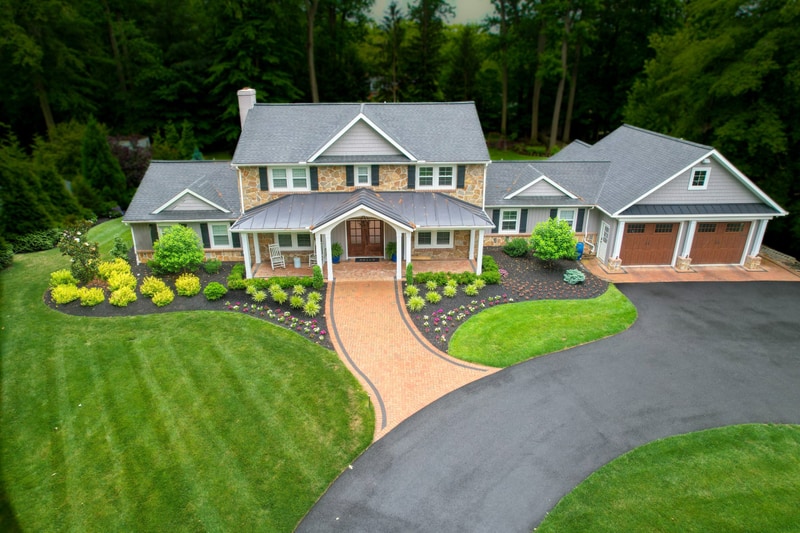 Aerial three-quarter view of the front of the North Wilmington home: the stone-and-cedar-shake facade sits centered, the curved tan paver driveway with charcoal Belgium-block edge sweeps in from below, foundation beds of mounded boxwoods and seasonal color trace the front, and a smooth lawn rolls down to the camera