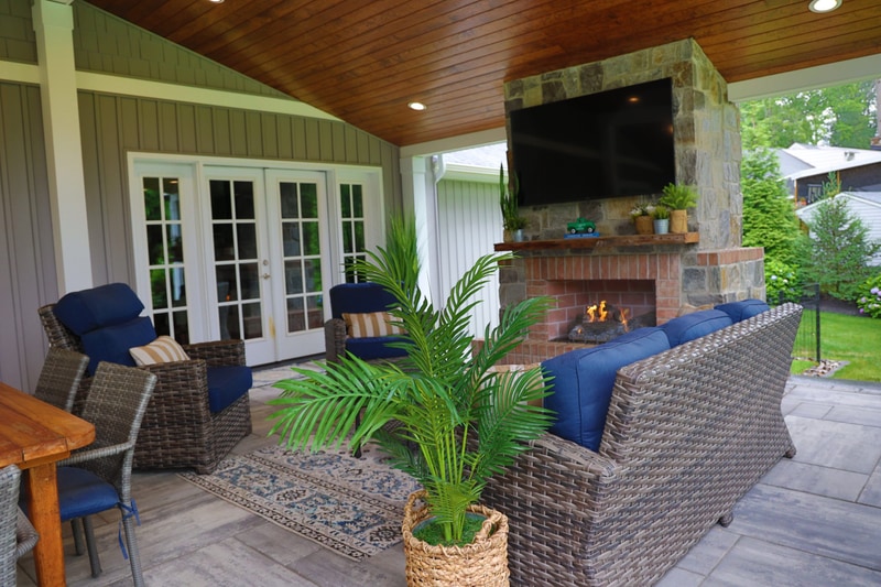 Inside the North Wilmington pavilion: a stone-faced fireplace with a mounted outdoor TV anchors the right side, a wicker sectional with navy cushions and patterned pillows wraps the seating area, a faux palm in a woven basket sits at center, the cathedral wood ceiling overhead, and french doors leading to the home at left