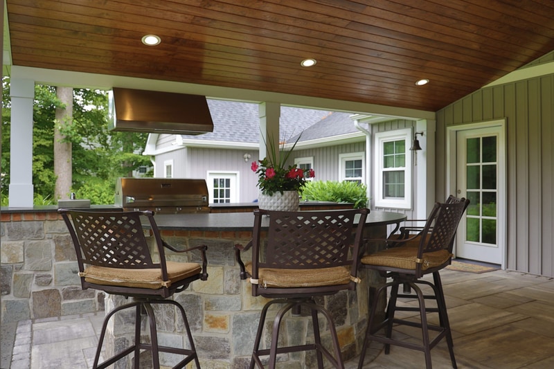 Wide view of the North Wilmington outdoor kitchen: four wicker swivel bar stools line the stone-faced bar, a stainless gas grill with copper-toned tile backsplash and a range hood are built into the rear wall, the cathedral wood ceiling overhead, and a vase of pink flowers on the bar