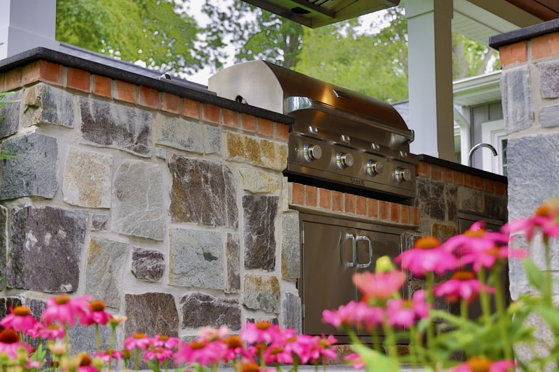 Close-up of the North Wilmington outdoor kitchen: the stone-faced bar with a stainless gas grill in focus, copper-toned tile backsplash, a small smoker on top of the grill, and a row of pink coneflower blooms soft-focus in the foreground