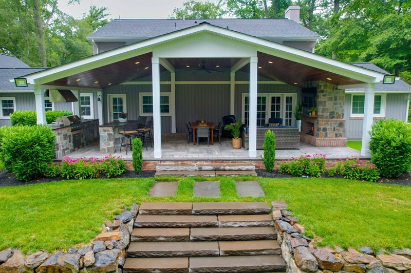Wide ground-level view of the North Wilmington back yard from the lawn: a sequence of large flagstone stepping-stone treads rises through the grass between dry-stack fieldstone retaining walls to the covered pavilion, with the dining and outdoor kitchen zones visible inside