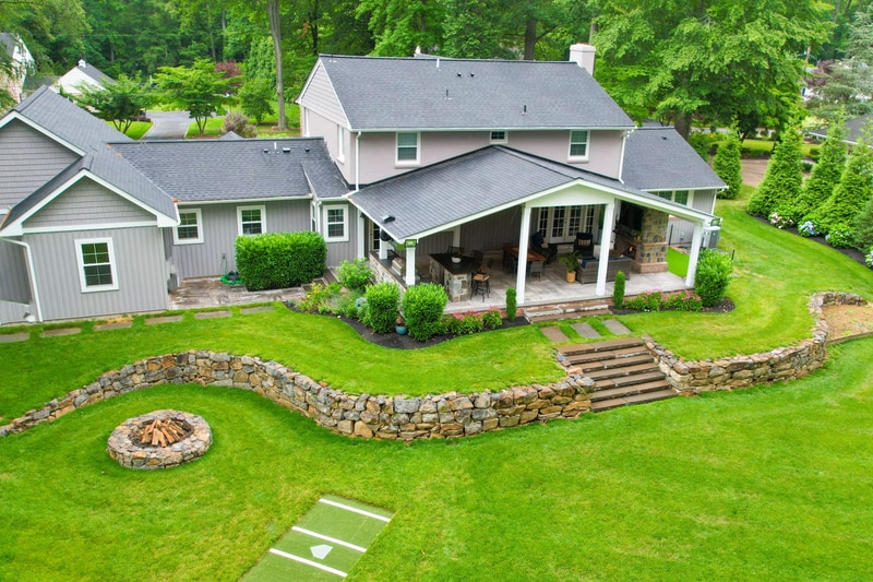 Aerial three-quarter view of the North Wilmington back yard: the covered timber pavilion at center, the curved dry-stack fieldstone retaining wall sweeping from left to right, hand-cut stone tread steps connecting the lawn to the patio level, and the round dry-stack fire pit visible in the lawn at lower left