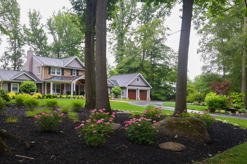 Wide street-level view of the North Wilmington property from across the lawn: pink mophead hydrangeas and a fieldstone foundation in the foreground, the home set among mature trees beyond, and the wing of the home with twin garage doors at right