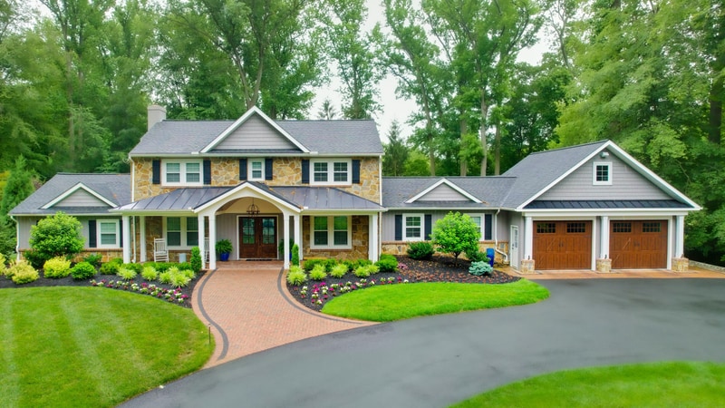 Front view of a North Wilmington Delaware home with a stone-and-cedar-shake facade: a curved tan paver driveway with charcoal Belgium-block edge sweeps from the lawn to the covered front porch, foundation beds of variegated liriope, mounded boxwoods, hydrangeas, and seasonal annuals running across the front, and mature trees framing the property