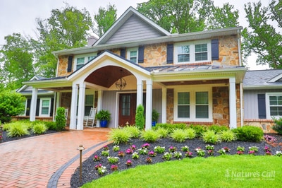 Ground-level view of a North Wilmington Delaware home: a stone-faced front elevation with a covered porch and tapered white columns, a brick paver walkway with charcoal Belgium-block edge sweeping in from the lawn, layered foundation plantings, hydrangeas in blue urns, and a band of pink and white annuals — Nature's Call landscape, hardscape, and outdoor living project.