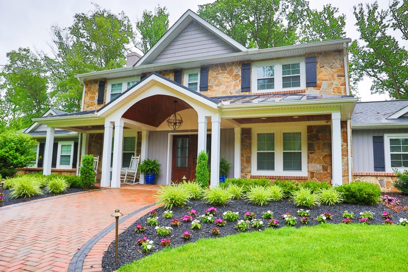 Front-elevation view of the North Wilmington home from across the lawn: the stone-and-shake facade with covered porch and double dark wood doors at center, the curved brick-paver walkway with charcoal Belgium-block edge approaching from below, foundation plantings of variegated liriope and pink-and-white annuals, and blue urn pots flanking the porch