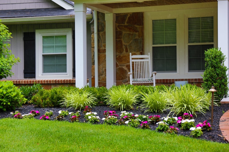 Detail of the North Wilmington front porch: a white wooden rocking chair sits on the covered porch, with the foundation bed below planted in variegated liriope and a border of pink and white impatiens with a path light at right