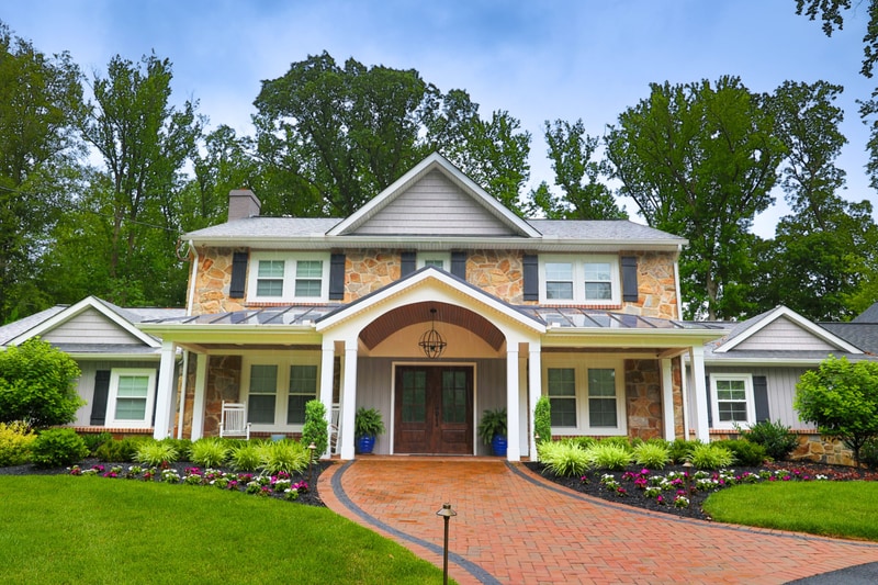 Frontal view of the North Wilmington front entry: the covered porch with white columns and lantern hangs above double dark wood front doors, blue ceramic urn pots with ornamental grasses flank the entry, and brick-paver walkway rolls in from below the foreground