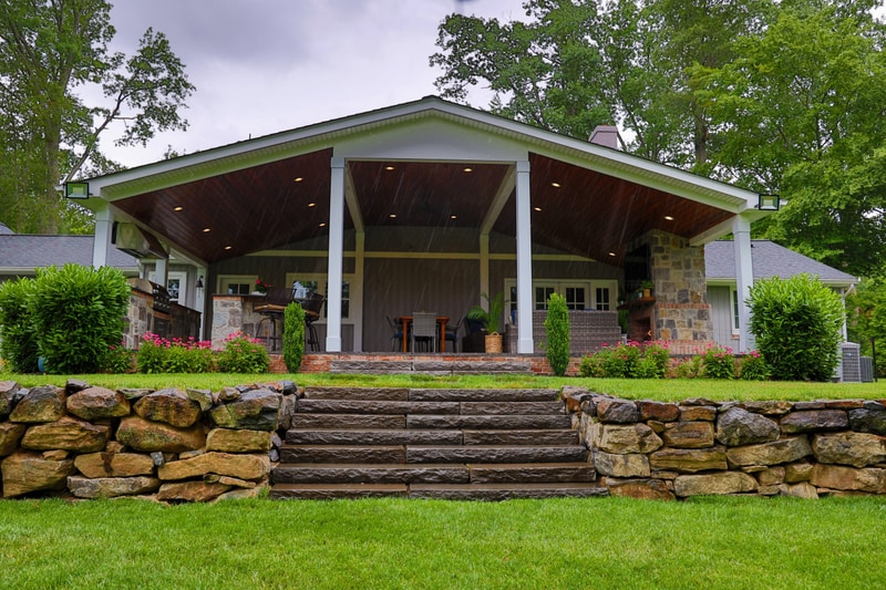 Ground-level view of the North Wilmington back yard: the dry-stack fieldstone retaining wall with hand-cut stone tread steps in the foreground rises to the covered timber pavilion with cathedral wood ceiling and recessed lighting, with the stone-faced outdoor kitchen and dining zone visible inside