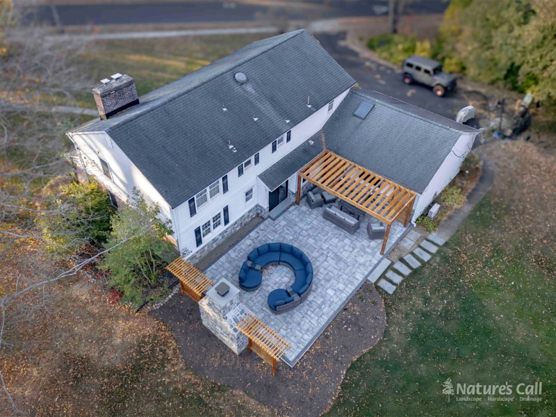Aerial dusk view of a North Wilmington Delaware property after Nature's Call rebuilt the back yard: a new gray paver patio with curved wicker sectional surrounds a stand-alone fieldstone outdoor fireplace, and a cedar pergola shades the patio against the home — Nature's Call hardscape and outdoor living project.