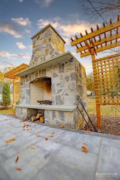 Golden-hour view of the stand-alone fieldstone outdoor fireplace with cedar pergolas flanking it, the firebox lit warm against a soft sunset sky, and stacked firewood and a small log holder at the base of the hearth