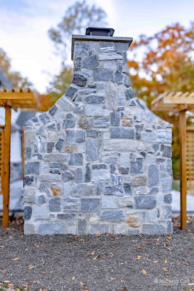 Head-on tight view of the stand-alone fieldstone outdoor fireplace: a tall fieldstone chimney with a metal cap rises against an autumn sky, the firebox and bluestone hearth at the base, and cedar lattice privacy screens visible to either side