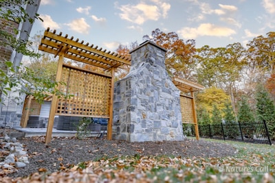 Side-angle view of the stand-alone fieldstone outdoor fireplace and the cedar lattice privacy screen behind it, with the home's chimney visible at right and a black aluminum fence and autumn trees beyond