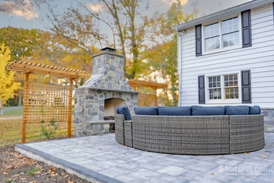 Evening view of the patio's seating zone: the curved wicker sectional with deep navy cushions wraps around the new fieldstone fireplace, the cedar pergola and lattice screen at left, and the home's stone-clad rear wall and double doors visible at right