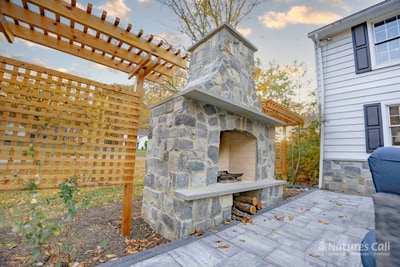 Side view of the cedar pergola and lattice privacy screen with the stand-alone fieldstone fireplace centered behind, and the gray paver patio extending to the right, framed by a strip of mulched bed and autumn lawn