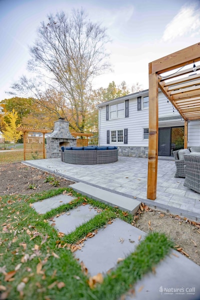 Bluestone stepping-stones running through the lawn up to the gray paver patio's edge, with the cedar pergola against the home at right, a stone-clad pier at the patio's near corner, and a fieldstone retaining edge along the patio's lawn side