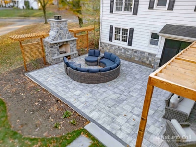 Three-quarter ground-level view at peak autumn color: the gray paver patio with curved wicker sectional and a small navy ottoman around the patio's seating zone, the stand-alone fieldstone outdoor fireplace at left framed by cedar lattice privacy screens, and the cedar pergola against the home at right