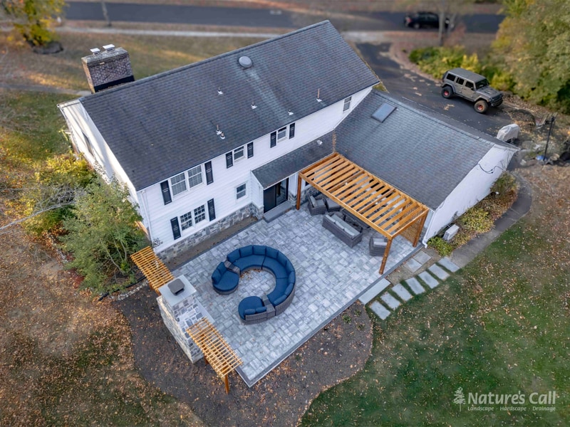 After. Aerial view of the same North Wilmington back yard rebuilt: a new gray paver patio with curved wicker sectional, a stand-alone fieldstone outdoor fireplace at one end, a natural cedar pergola against the home, lattice privacy screens flanking the fireplace, and the old stockade fence removed in favor of an open lawn